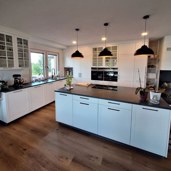 Eine moderne Küche mit weißen Schränken, schwarzen Arbeitsflächen und hängenden Lampen. Die Küche hat eine zentrale Insel mit Herd und einem großen Fenster.