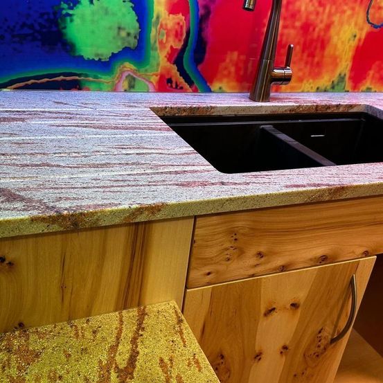 Eine Küchenspüle mit Granitarbeitsplatte, einem Wasserhahn und farbiger abstrakter Kunst an der Wand. Die Arbeitsplatte hat rote Streifen, während die Schränke aus Holz sind.