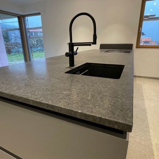 Eine moderne Küche mit Granitarbeitsplatte, einem schlanken schwarzen Wasserhahn und einem großen Fenster mit Blick nach draußen.