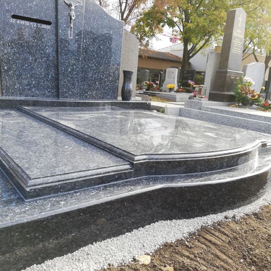 Ein Granitgrabstein mit einem Kreuz und einer Vase, in einem Friedhof gelegen. Dahinter sind Bäume und ein Gebäude zu sehen. Der Grabstein hat eine glänzende Oberfläche.