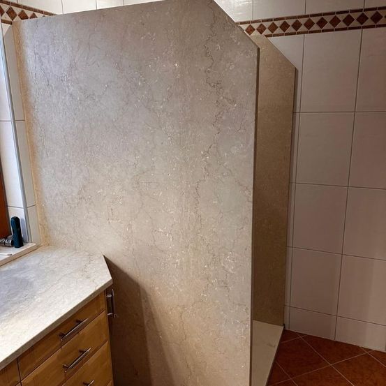 Ein Badezimmer mit einer Marmorarbeitsplatte, Holzschubladen und einer Duschkabine mit einer Marmorwand. Die Wand hat einen braunen Rand und einen weißen Fliesenrand.