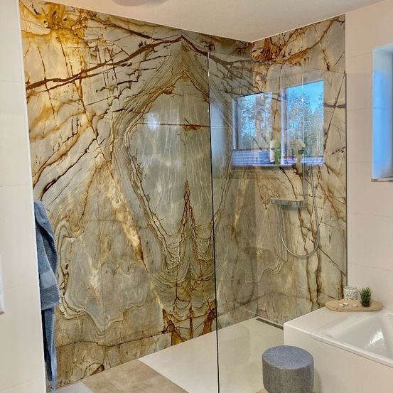 Ein modernes Badezimmer mit einer Glasdusche und Marmorwänden. Ein blaues Handtuch hängt links, und eine Pflanze steht auf der Badewanne. Das Fenster zeigt Bäume.