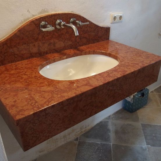 Ein Marmorarbeitsplatte mit einem weißen Waschbecken und Wasserhahn, an einer weißen Wand montiert. Eine Steckdose ist an der Wand über der Arbeitsplatte montiert. Ein blauer Plastikbehälter steht auf dem Boden.