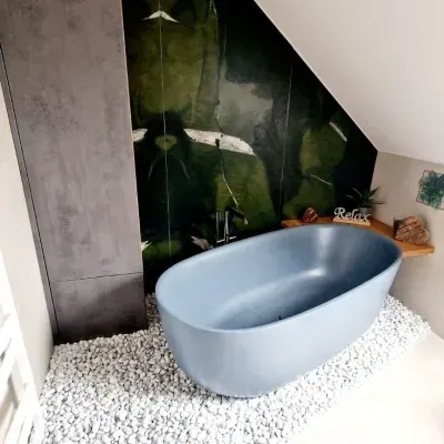 Eine blaue Badewanne steht auf einem weißen kiesbedeckten Boden, mit einer grauen Wand und einer grünen Blattwand dahinter. Es gibt einen Wasserhahn an der Wand.