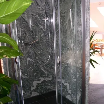 Eine moderne Duschkabine mit einer Glastür und schwarzem Fliesenboden. Sie verfügt über einen silbernen Duschkopf und einen silbernen Griff, vor einer granitartigen Wand.