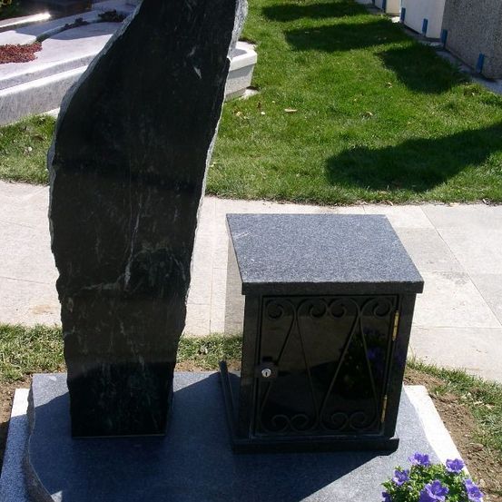 Ein großes schwarzes Granitmonument steht auf einem Betonsockel. Daneben befindet sich ein kleinerer schwarzer Kasten mit einem Metalldesign. Der Kasten hat einen Schloss und ist von einem kleinen Strauß lila Blumen umgeben.