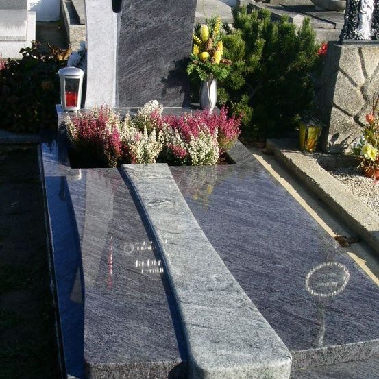 Ein Friedhof mit einem Grabstein auf einer Betonplattform, geschmückt mit Blumen, einer Kerze und einer Vase, umgeben von Grünpflanzen und Pflanzen.