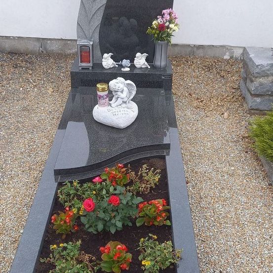 Ein grauer Grabstein mit Blumenvase und kleinen Statuen. Vor dem Grabstein befindet sich ein Blumenbeet mit Rosen und anderen Blumen.