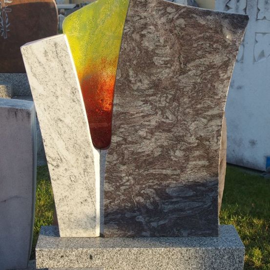 Ein Granitgrabstein mit farbiger Glaseinlage wird im Freien ausgestellt. Das Glas zeigt leuchtende Rot-, Gelb- und Grüntöne.