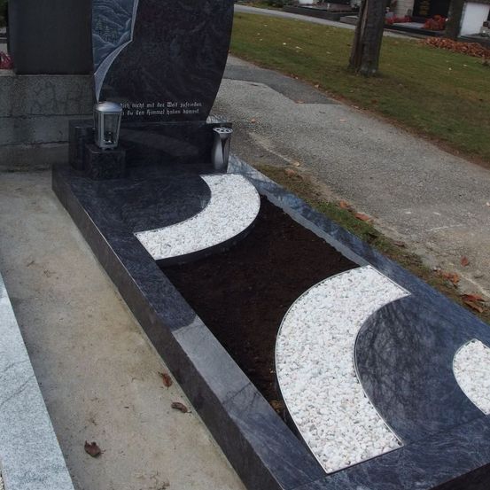 Ein Grab mit einem schwarzen Granit-Grabstein und zwei Lampen, einem Weg mit Rasen an den Seiten und verstreuten Blättern.