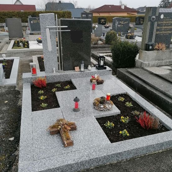 Ein grauer Grabstein auf einem Friedhof mit einem Holzkreuz und Blumen. Mehrere rote Laternen und Kerzen sind auf dem Boden platziert.
