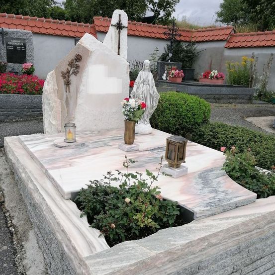 Ein Marmorgrab mit einer Statue der Maria, einem Kreuz und Blumen in Vasen. In der Nähe sind Topfpflanzen und ein Gebäude mit rotem Dach.