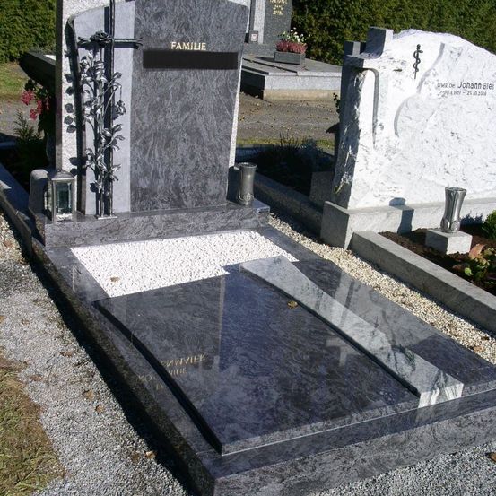 Ein grauer Granit-Familiengrabstein mit einem Kreuz, zwei Vasen und einer Metalllaterne. Die Inschrift lautet 'FAMILIE'. Daneben steht ein weißer Grabstein, der teilweise verdeckt ist.