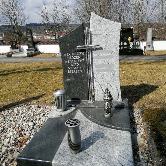 Ein Steindenkmal mit einem Kreuz und einer Statue steht auf einem Friedhof. Die Inschrift lautet: 'Wer in deinem Herzen lebt, wird nie sterben.' Vorne steht eine Lampe.