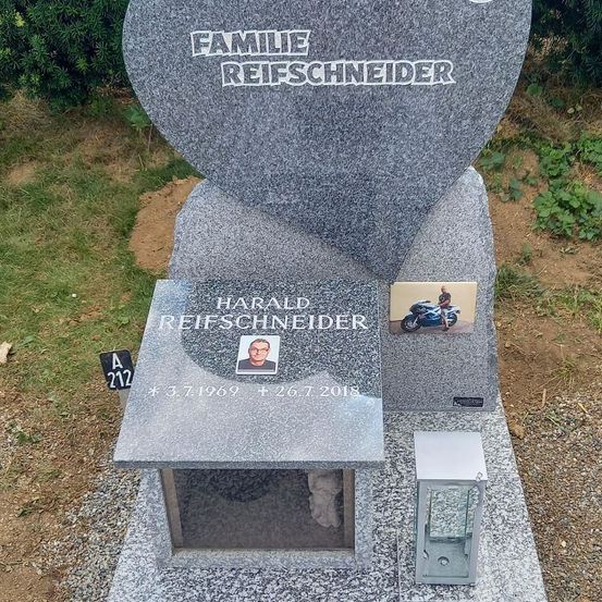 Ein graues Grabmal für die Familie Reifschneider mit einem Foto von Harald Reifschneider und einem Motorradbild. Das Grabmal befindet sich auf einem Friedhof mit einem mit kleinen Steinen und Gras bedeckten Boden.