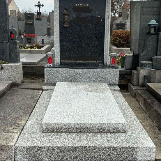 Ein Friedhof mit mehreren Grabsteinen, einer mit einer schwarzen Plakette, auf der das Wort Familie steht. Oben befindet sich eine Statue einer Frau. Auf beiden Seiten der Plakette stehen rote Kerzen.