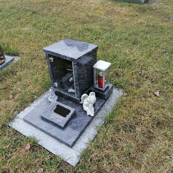 Ein Grabstein auf einer Rasenfläche mit einer Glasbox, in der eine Kerze steht, einer kleinen Engelsstatue und einem Kreuz. Der Sockel ist grau, und es gibt eine Tafel mit gravierten Texten.