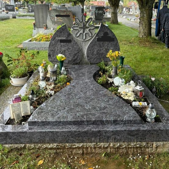 Ein Friedhof mit einem grauen Grabstein, geschmückt mit Blumen und einem Buch. Es gibt auch andere Grabsteine und Bäume in der Umgebung.
