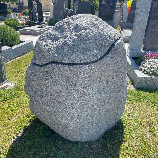 Eine große Steinskulptur mit einer rissigen Oberfläche steht auf einer Rasenfläche, möglicherweise einem Friedhof.