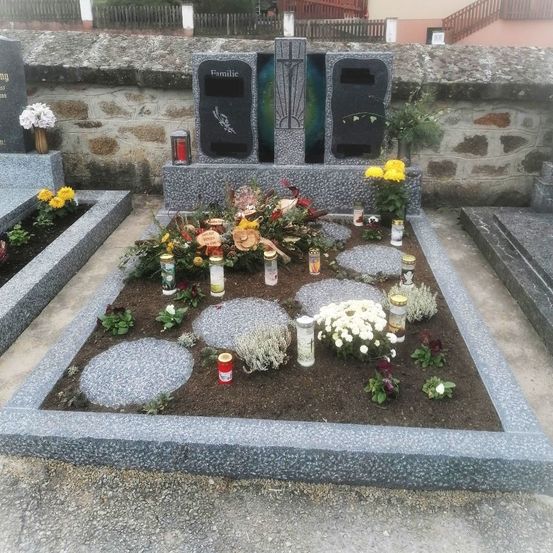 Ein Friedhof mit einer grauen Steinstruktur hat ein großes Kreuz, eine Tafel mit der Aufschrift 'Familie' und ein Grab, das mit Blumen, Kerzen und Steinen geschmückt ist. Das Grab ist von anderen Grabsteinen umgeben und von einer Steinmauer umschlossen.