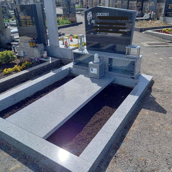 Ein graues Granit-Familiengrab auf einem Friedhof. Das Grab hat einen offenen Raum mit Erde und ist von Blumen und Laternen umgeben.
