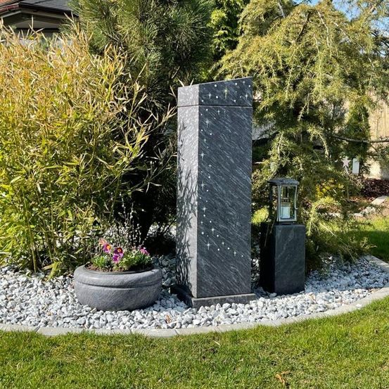 Ein Garten mit einem hohen, sternverzierten Steinmonument. In der Nähe stehen eine Laterne und ein Blumentopf auf Kies.