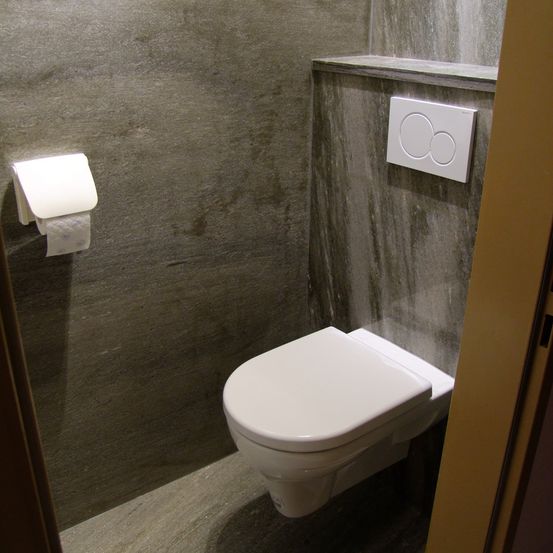 Ein modernes Badezimmer mit einer grauen Betonwand, einer weißen Toilette und einem weißen Toilettenpapierhalter über der Toilette. Die Wand hat neben der Toilette einen Schalter.