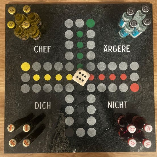 Ein Brettspiel wird mit Würfeln, Chips und Flaschen angezeigt. Deutsche Wörter sind Chef, Dich, Argere, Nicht. Flaschen mit Etiketten sind um das Brett herum angeordnet.