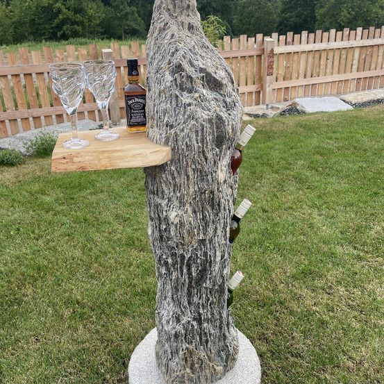 Ein Baumstamm mit einer Flasche Jack Daniel's Whiskey, zwei Weingläsern und mehreren Weinflaschen, eingebettet in ihn, auf einem Betonsockel.