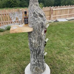 Ein Baumstamm mit einer Flasche Jack Daniel's Whiskey, zwei Weingläsern und mehreren Weinflaschen, eingebettet in ihn, auf einem Betonsockel.