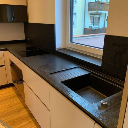 Eine moderne Küche mit einer schwarzen Granitarbeitsplatte, zwei Spülen und einem Fenster mit Blick auf ein Gebäude mit Balkon.