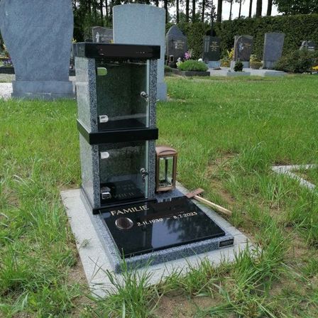 Ein schwarzer Grabstein mit einer Glasbox oben steht auf einem Friedhof mit grünem Gras. Der Grabstein hat das Wort 'FAMILIE' und ein Datum. Eine Laterne und ein Hammer sind auf dem Grabstein platziert. Hinter dem Grabstein befinden sich andere Grabsteine mit Pflanzen und Blumen.