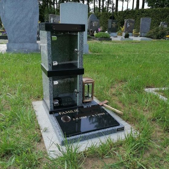 Ein schwarzer Grabstein mit einer Glasbox oben steht auf einem Friedhof mit grünem Gras. Der Grabstein hat das Wort 'FAMILIE' und ein Datum. Eine Laterne und ein Hammer sind auf dem Grabstein platziert. Hinter dem Grabstein befinden sich andere Grabsteine mit Pflanzen und Blumen.