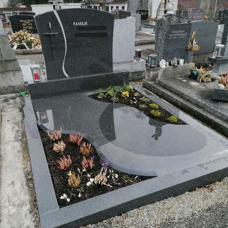 Ein Friedhof mit einem grauen Grabstein, auf dem 'FAMILIE' steht. Er ist von Blumen, Kerzen und Laternen umgeben. Der Grabstein hat ein Kreuz und ein geschwungenes Design.