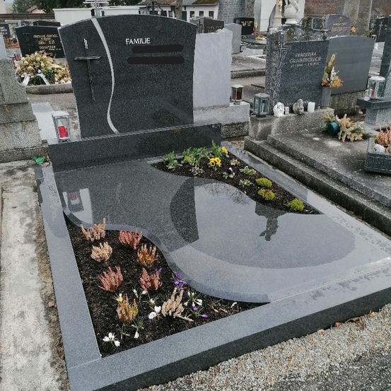 Ein Friedhof mit einem grauen Grabstein, auf dem 'FAMILIE' steht. Er ist von Blumen, Kerzen und Laternen umgeben. Der Grabstein hat ein Kreuz und ein geschwungenes Design.