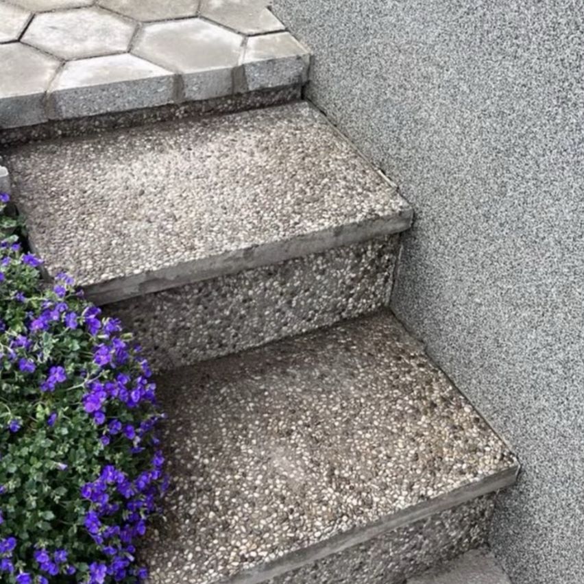A close-up view of three concrete steps with a textured surface, adjacent to a wall with a small plant with purple flowers on the left.