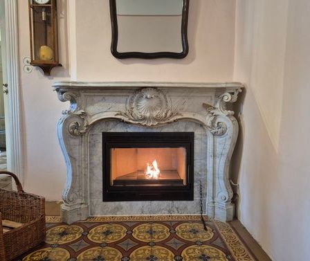 Ein Zimmer mit einem Kamin mit einem verzierten Marmorsims und einem brennenden Feuer, begleitet von einem Korb mit Holz und einer Wanduhr.