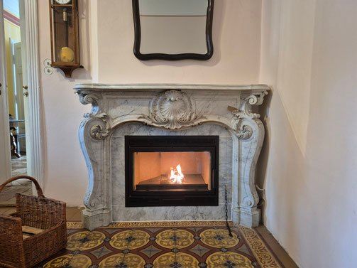 Ein Zimmer mit einem Kamin mit einem verzierten Marmorsims und einem brennenden Feuer, begleitet von einem Korb mit Holz und einer Wanduhr.