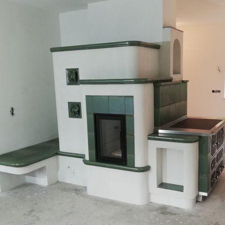 Ein Innenraum mit einem modernen Kamin, grünen Fliesen und einer Bank. Der Raum ist unfertig mit sichtbaren Betonböden und weißen Wänden.