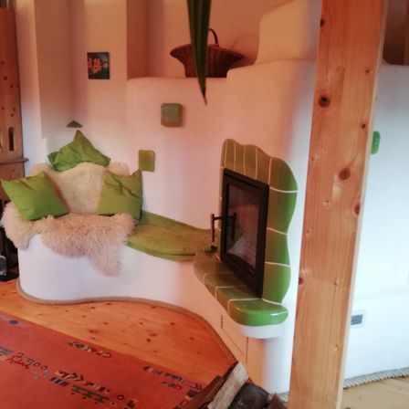 Eine gemütliche Ecke mit einer weißen und grünen Bank, grünen Kissen und einem Kamin mit grüner Fliesenumrandung. Ein Korb steht auf dem Kamin.