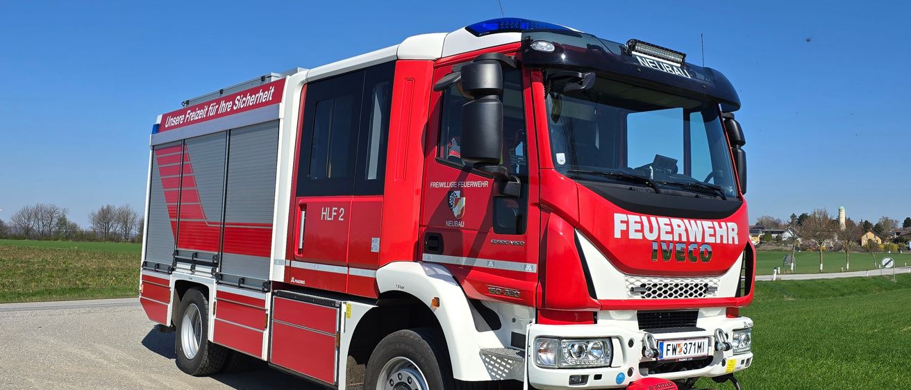 Ein roter und weißer Feuerwehrwagen steht auf einer Straße mit klarem blauem Himmel im Hintergrund. Der Wagen trägt die Aufschrift 'FEUERWEHR IVECO' auf der Vorderseite und ein Nummernschild mit der Aufschrift 'FW 371 MI'.