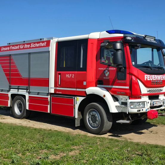 Ein roter und weißer Feuerwehrwagen mit der Aufschrift 'Unsere Freizeit für Ihre Sicherheit' auf einem Feldweg geparkt. Das Kennzeichen lautet 'FW 37TM1'.