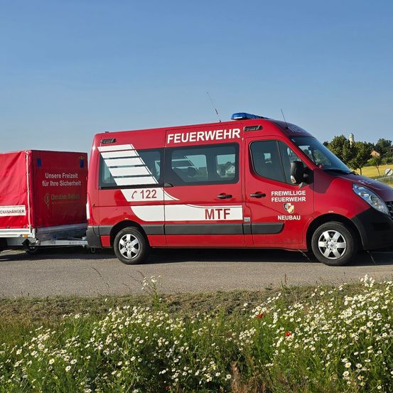 Ein roter Feuerwehrwagen mit Anhänger auf einer Straße, umgeben von Wildblumen. Der Wagen trägt 'FEUERWEHR' und 'FREIWILLIIGE FEUERWEHR' an der Seite.