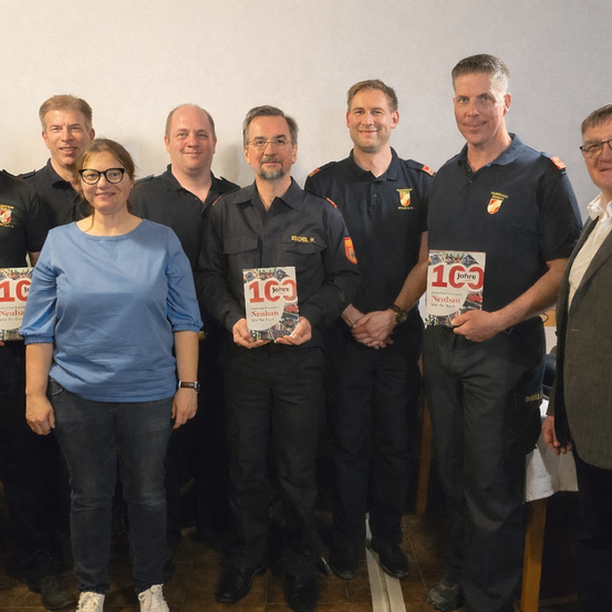 Eine Gruppe von sieben Personen steht in einer Reihe und hält Bücher in den Händen. Sie sind alle in Uniform gekleidet. Die Personen in der Mitte lächeln und posieren für ein Foto. Die Person ganz rechts trägt einen Anzug.