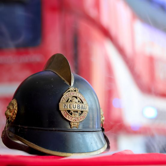 Ein vintage schwarzer Feuerwehrhelm mit goldenen Akzenten und einem goldenen Emblem mit der Aufschrift 'NEUBAU' wird vor einem unscharfen roten Feuerwehrwagen-Hintergrund präsentiert.