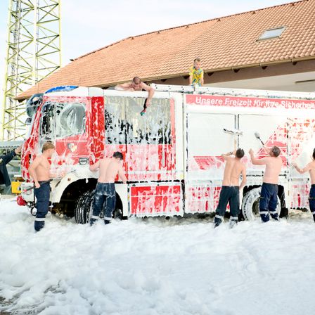 Fünf Feuerwehrleute waschen einen Feuerwehrwagen mit Schaum vor einem Gebäude. Ein Junge sitzt oben auf dem Wagen.