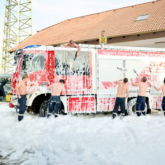Fünf Feuerwehrleute waschen einen Feuerwehrwagen mit Schaum vor einem Gebäude. Ein Junge sitzt oben auf dem Wagen.