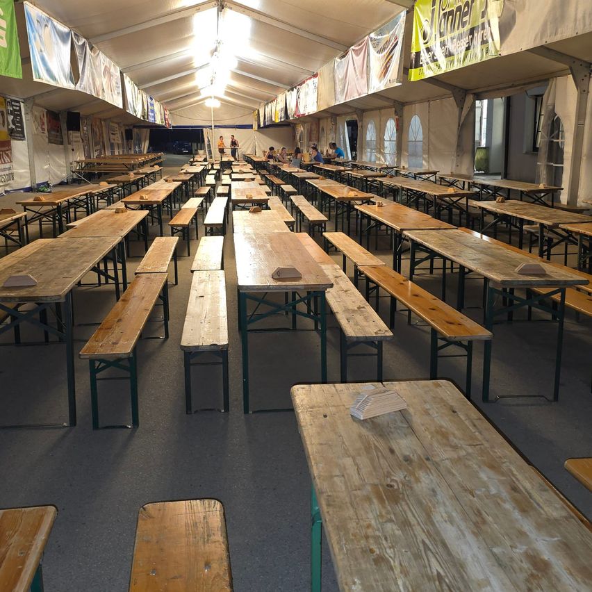 Ein leerer Zeltraum mit Reihen von Holztischen und Bänken, beleuchtet von Deckenlampen. Banner hängen von der Decke, und einige Leute sitzen in der Ferne an Tischen.