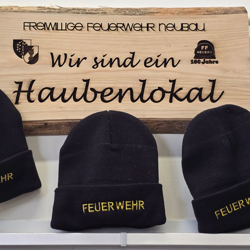 Drei schwarze Mützen mit gelbem Text 'Feuerwehr' werden vor einem Holzschild präsentiert, auf dem 'Fremwillige Feuerwehr Neubau' und 'Wir sind ein Haubenlokal' steht. Das Schild zeigt auch ein Logo und den Text '100 Jahre'.