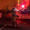 Eine Person in einem gehörnten Kostüm hält eine brennende Fackel in der Mitte einer Straße, umgeben von Rauch und einer Menge von Menschen. Hinter ihm sind Banner und Schilder sichtbar, darunter eines mit der Aufschrift 'Tischlerhans'. Im Hintergrund sind Gebäude und Straßenlaternen zu sehen.